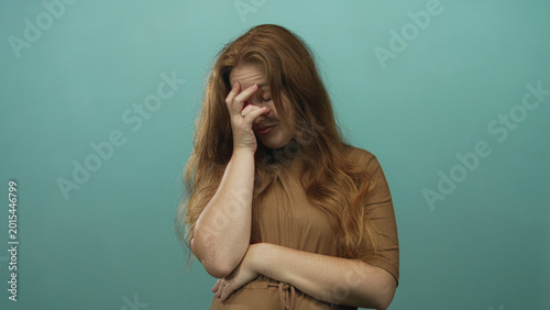 Young redhead woman with hand covers face and crosses arms in studio with teal backdrop; embarrassment.