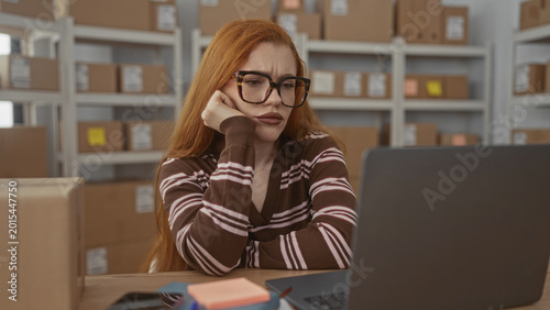 Foto Redhead woman rests chin on hand while staring at laptop in a warehouse building filled with cardboard boxes and shelving; frustration fatigue