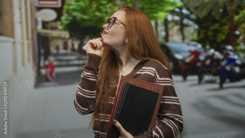 Foto Young redhead woman holding a hardcover book with glasses, hand to chin gesture and slight smile while standing near parked scooters and cars on a street; thoughtful curiosity