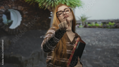 Photos Young redhead woman points index finger at camera while clutching a hardcover book in front of an urban building rooftop; confidence attitude