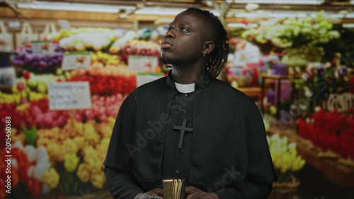 Bild auf Leinwand Young priest man holding book with visible hands and clerical collar in flower market stalls; faith serenity