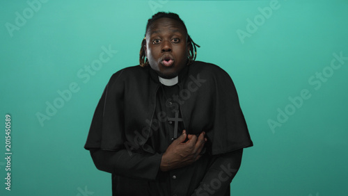 Bild auf Leinwand Young priest man holding hand to chest with mouth open in studio against teal backdrop; devotion prayer faith