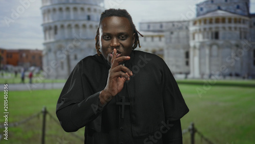 Fotografie Man in clerical robes wearing a prominent cross necklace, finger to lips for silence in building at the leaning tower of pisa plaza; silence reflection