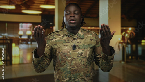 Canvas Print Man in camouflage uniform raises palms and gestures with dog tags in a hotel building, eyes closed while speaking softly; duty resolve resilience