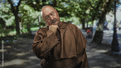 Bild auf Leinwand Man monk in brown robe with hand to chin and visible wooden cross necklace, standing under trees on a sunlit park path; prayerful reflection serenity