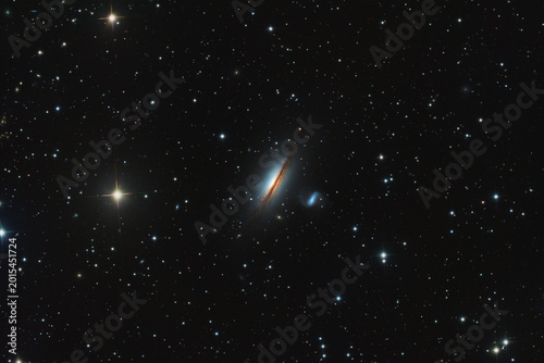 A astrophotography image of the edge-on lenticular galaxy NGC 5078 featuring a prominent dark dust lane and a nearby companion galaxy set against a dense field of distant stars. Telescope live.