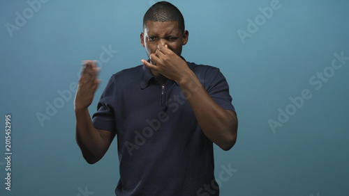 Valokuva Man pinching nose with hand in studio, wearing navy polo shirt, short hair, grimacing and raising the other hand in front of a plain blue backdrop; disgust reaction