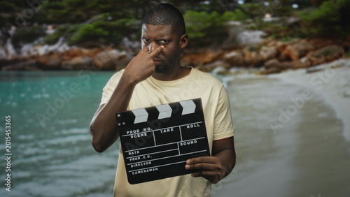 Fotografie Man holding clapperboard pinching nose on a studio set with beach backdrop, staring at camera with a sneer; disgust satire