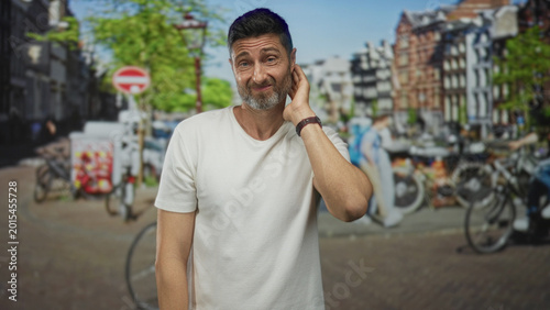 Foto Man touching neck with hand and wry half smile beside bicycles by a canal on a street in amsterdam; awkwardness uncertainty