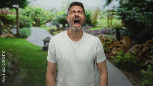 Man shouting with open mouth on forest path in green park, visible mouth and open jaw, white shirt visible; anger release solitude.