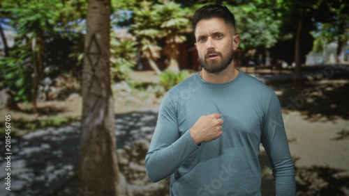Vászonkép Hispanic man with beard stands in a shaded forest, places hand over chest then lifts both palms outward in a pleading gesture; apology