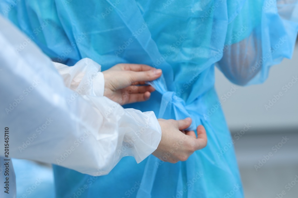 Fototapeta premium Medical assistant putting isolation gown on doctor indoors, closeup