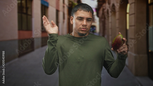 Quadro em tela Man grimacing while holding and inspecting a mango in a narrow street archway by buildings, pointing hand toward fruit; disgust caution
