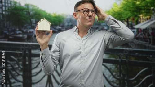 Bild auf Leinwand Man holding tiny wooden house while scratching head on street bridge with slight squint and eyeglasses visible; uncertainty mortgage decision