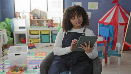 Konstfotografi Woman teacher tapping tablet while seated in a colorful school classroom with toys and play tent; focus lesson planning