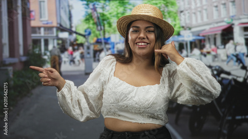 Photos Woman in straw hat points finger left and makes phone gesture with thumb to ear, crop top exposing midriff on street; confident casual travel
