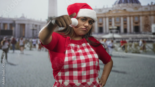 Bilde på lerret Woman in santa hat and red check apron gives thumbs up at camera in a crowded street by a monumental plaza with colonnades and obelisk; festive