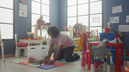 Fotografía Man kneels picking up toy blocks on foam mat and holds plastic brick near backpack in kindergarten building; nurturing calm