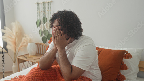 Schilderij op canvas Man sitting on bed covering face with hands, orange blanket and pillow and bedside vase visible inside building; fatigue introspection