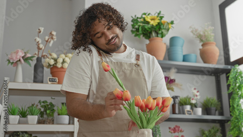 Man arranging orange tulips with phone held at shoulder while adjusting stems in apron inside a flower shop building; calm focus.