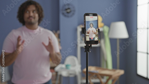 Konstfotografi Man gesturing with hands beside wooden cradle and smartphone on tripod in studio, speaking to camera; nurturing care