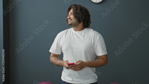 Φωτογραφία Man tapping red smartphone with fingers while standing in studio gym, wearing white tee and focused on screen; calm attentive focus