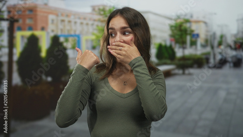 Canvas-taulu Young hispanic woman wearing green top and heart necklace, covering mouth with hand and pointing thumb on street; surprise