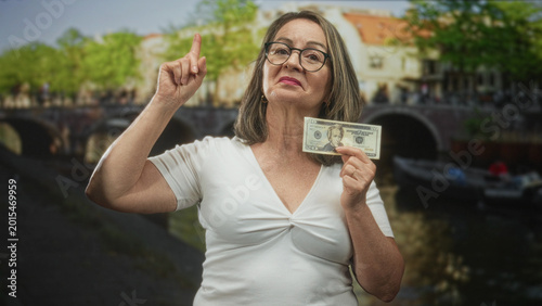 Foto Hispanic senior woman points finger up while holding a twenty banknote by a canal bridge on a street; confidence savings