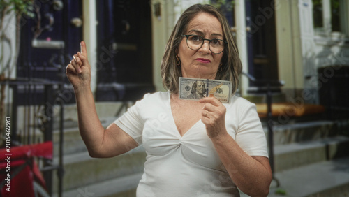Cuadro en lienzo Woman grey hair holds a hundred dollar banknote and points finger upward at a building stoop with raised arm; financial frustration concern