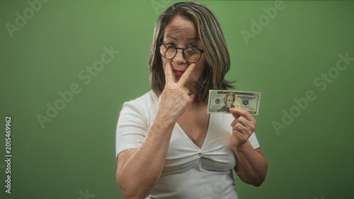 Tableau sur toile Hispanic senior woman wearing glasses and a white top holds a twenty dollar banknote and makes a v sign with fingers near her nose in studio; assertive warning