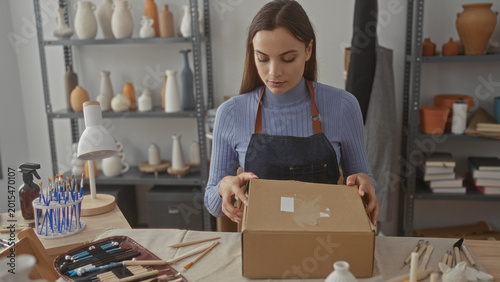 Photos Woman opening a cardboard box on a wooden worktable in studio wearing an apron and handling tape surrounded by pottery on shelves; concentration craft packaging