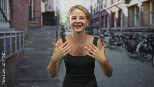 Tableau sur toile Blond woman touching chest with both hands showing bare shoulders on street lined with bicycles; joyful confidence