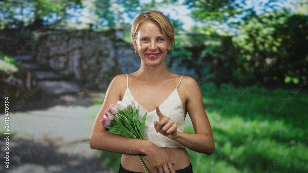 Naklejka premium Blond woman smiling, holding pink tulips and pointing finger along a sunlit forest path; joy optimism warmth.