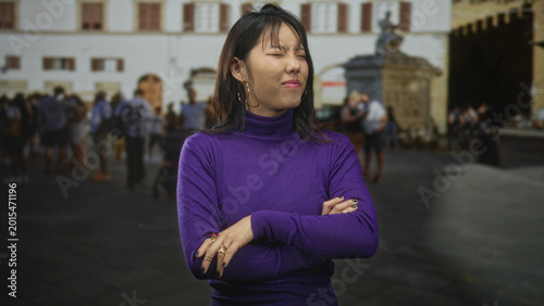 Foto Woman asian with arms crossed and squinting face wearing purple sweater on street by fountain and tourist crowd in front of historic building; annoyance rejection