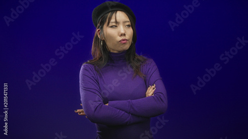 Woman in purple turtleneck wearing beret with crossed arms and one thumb up gesture in studio; wry amusement.