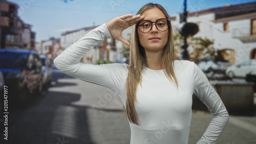 Fotografija Young hispanic woman wearing glasses salutes with hand to forehead while standing on a sunlit street lined with parked cars and buildings; confidence focus