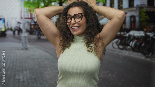 Fotografija Woman in green top wearing glasses making ear gesture with hands on a bustling city street; playful mood