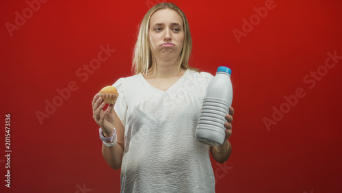 Photography Woman holding muffin and large milk bottle, pouting lips, bare hands and white blouse against red studio backdrop; indecision