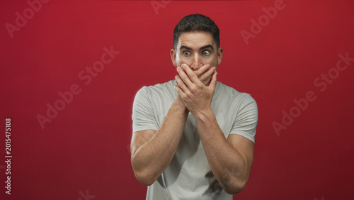 Tableau sur toile Young hispanic man in gray tshirt with hands covering mouth in red studio; surprise secrecy hush secret