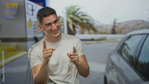Cuadro en lienzo Man smiling and pointing finger toward camera at petrol pump beside silver car on street; confidence playful
