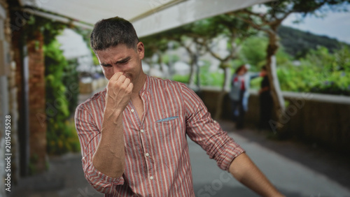 Foto Man pinching nose on street promenade near cafe, hand over nostrils and eyes squinted; disgust odor avoidance