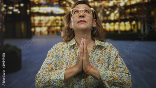 Bild auf Leinwand Woman wearing floral blouse and glasses with clasped hands praying on an illuminated street in front of building lights; hope prayer