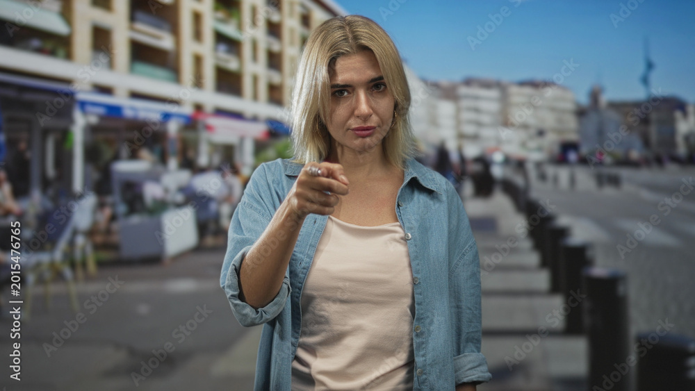 Fototapeta premium Woman pointing finger at camera on a crowded street wearing denim shirt and beige tank top; accusation.
