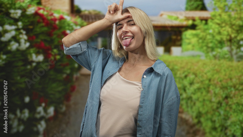 Fotografija Woman with blonde hair in denim shirt makes l sign on forehead and sticks tongue out toward camera beside residential building entrance; playful mischief
