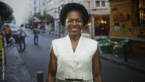Obraz na płótnie Woman smiling in white sleeveless vest standing centered on a busy street with green outdoor chairs and a bollard visible; urban confidence