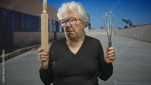 Foto Elderly woman holds a rolling pin and metal whisk in her hands, frowning and pointing slightly in front of a building with windows, wearing colorful glasses and red nails; grumpy humor