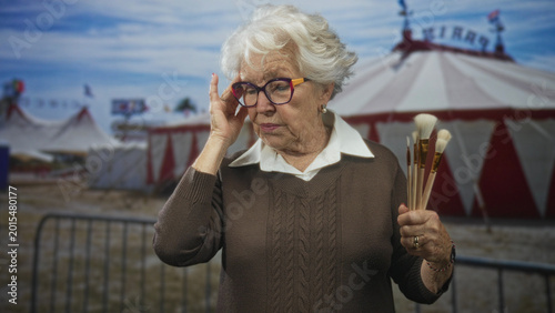 Tableau sur toile Elderly woman holding paintbrushes touches eyeglasses on street in front of circus tent, slight frown and thoughtful gaze; nostalgia calm