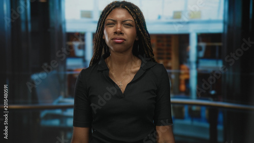 Foto Woman squints eyes and presses lips, wearing a cross necklace and braided hair and a black collared shirt in cruise ship building interior; seasick discomfort