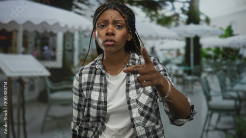 Foto Woman with arms crossed on outdoor restaurant terrace wearing plaid shirt and white tee, braided hair and stern expression; frustration refusal