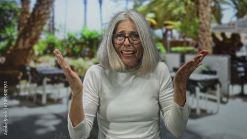 Cuadro en lienzo Woman raising both hands with pleading palms at outdoor restaurant terrace, grey hair and glasses; frustration confusion